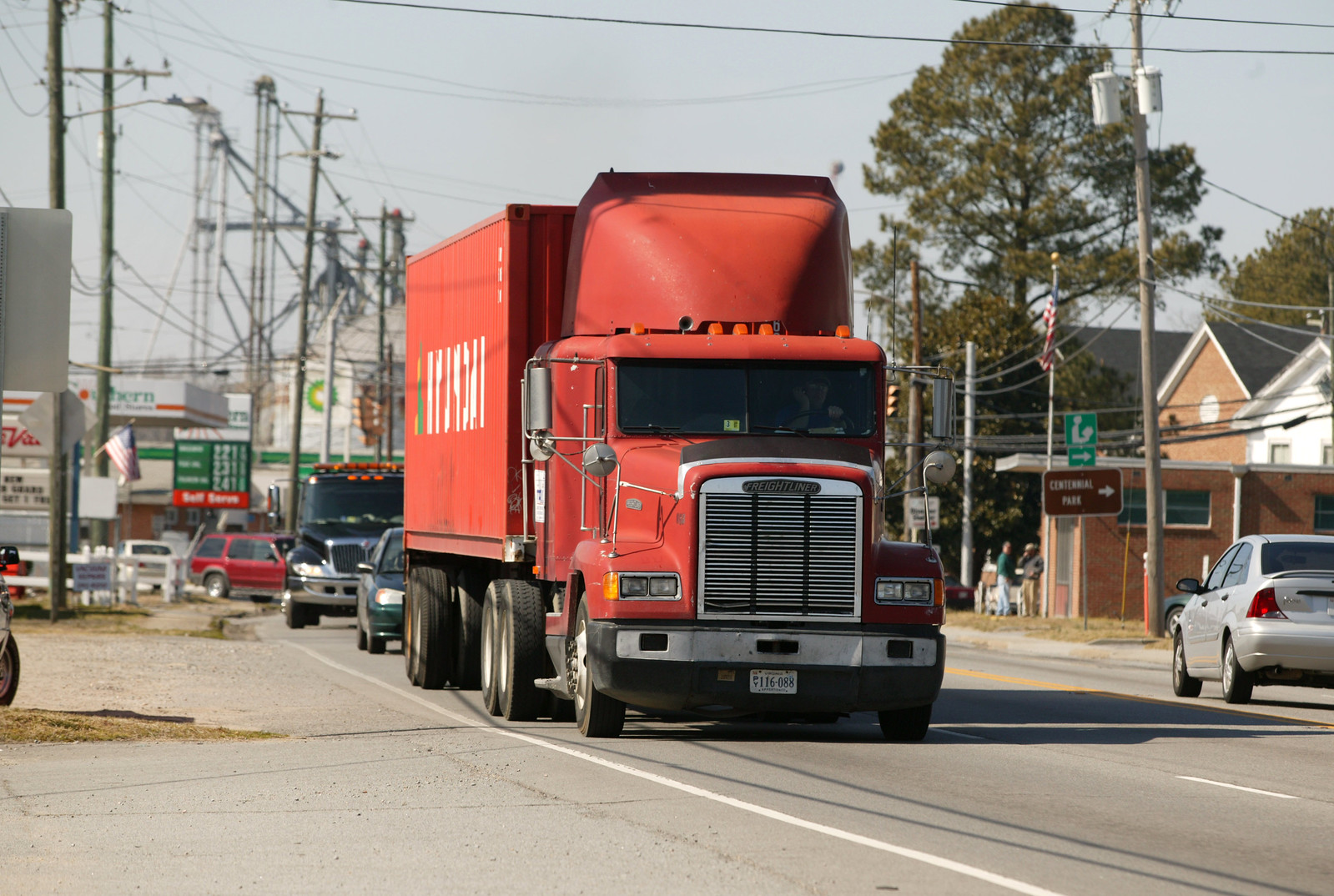 A red container truck leaving the port and driving through streets near houses and gas stations.